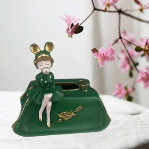 May include: A decorative green tissue box holder featuring a figurine of a girl wearing a green dress and a mouse-ear hat. The holder has gold accents and a heart-shaped design with the words "Love You". Pink flowers are in the background.