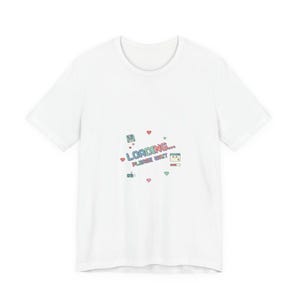 Könnte beinhalten: Weißes T-Shirt mit einem farbenfrohen Grafikdesign. Das Design zeigt die Worte "LOADING... PLEASE WAIT" in einer verspielten Schriftart, pixelige Herzen, einen Gamecontroller und ein Computerbildschirm-Symbol.