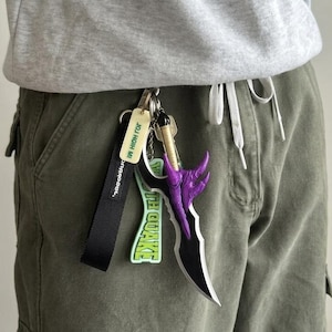 Puede incluir: Un llavero con un dije de daga negra y morada, un cordón negro con texto blanco y una etiqueta verde con el texto "THE QUAKE". El llavero está sujeto a un par de llaves.