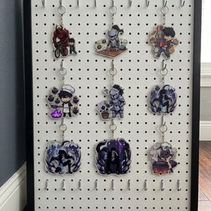 May include: A black framed pegboard displaying nine anime-style acrylic keychains. Each keychain features a different character design, with vibrant colors and detailed artwork. The keychains are suspended from metal hooks, creating a visually appealing display.