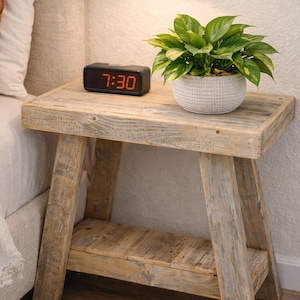 May include: A rustic wooden nightstand with a small black digital clock displaying 7:30. A potted plant with green leaves sits on top. A gold-toned lamp is mounted on the wall above.
