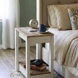 May include: A rustic, white-painted wooden side table with a distressed, natural wood top. A gold clock, a blue mug, and a wireless charger sit on top. Below, a shelf holds a book and headphones. The table is next to a bed.