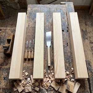 May include: A woodworking scene featuring four unfinished wooden beams, a hand saw, chisels, and a wooden plane. Wood shavings are scattered on the work surface, indicating recent work. The tools and beams suggest a carpentry or woodworking project.