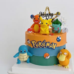Puede incluir: Un pastel de varios niveles con glaseado naranja, azul y verde que presenta el logotipo azul de Pokémon. El pastel está decorado con figuras de plástico de Charmander, Pikachu, Bulbasaur, Squirtle y Psyduck, junto con detalles de Poké Ball y un adorno de Feliz Cumpleaños.