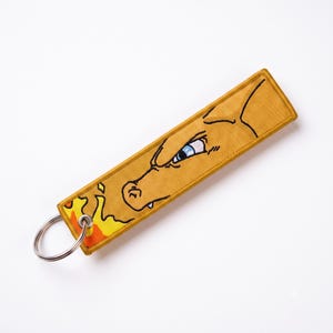 May include: A rectangular mustard yellow fabric keychain featuring an embroidered profile of a dragon-like creature with a blue eye and a small white tooth. The design includes a stylized orange and yellow flame graphic and a silver metal ring.