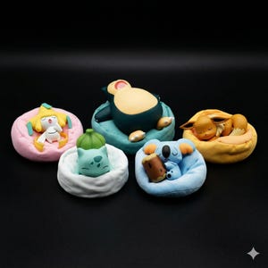 Puede incluir: Cinco figuras coloridas de personajes durmientes, cada uno descansando sobre un cojín en miniatura. Los personajes incluyen una figura amarilla y blanca sobre rosa, una figura azul y beige sobre turquesa, una figura verde sobre blanco, un koala sobre azul y un zorro sobre amarillo.