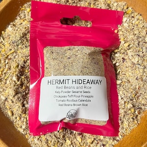 May include: A bright pink pouch labeled "HERMIT HIDEAWAY" containing a blend of ingredients, including red beans, rice, and seeds. The pouch is set in a wooden bowl filled with similar dry ingredients. The label lists the ingredients.