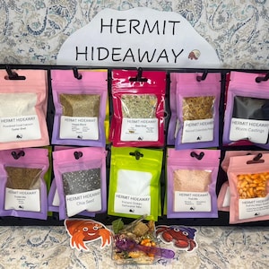 May include: A collection of small, clear plastic bags filled with various hermit crab food items. The bags are labeled with the text "HERMIT HIDEAWAY" and the type of food inside, such as "Worm Castings", "Chia Seed", and "Instant Ocean Saltwater Mix".