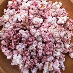May include: A close-up shot of a wooden bowl filled with gourmet popcorn. The popcorn kernels are a mix of white and deep red, with a speckled pattern. The kernels are popped and appear fluffy, with some showing hints of yellow.