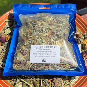 May include: A blue resealable bag filled with a mix of dried herbs and flowers. The bag is labeled "HERMIT HIDEAWAY Walk in the Field" and lists the ingredients: Timothy Hay, Alfalfa Hay, Peas, Hibiscus Flower, Marigold Flower, Rose Petals, Nettle Leaves, Birch Leaves, Raspberry Leaves, Blue Cornflowers, and Chamomile Flowers.