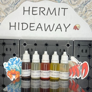 May include: Six small bottles of essential oil labeled with different scents: Sesame Oil, Sunflower, Chili Oil, Walnut Oil, and Safflower. The bottles are arranged in a row on a black surface. The text "HERMIT HIDEAWAY" is visible at the top of the image.