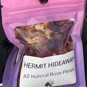May include: A pink and purple bag containing dried rose petals. The bag has a clear window displaying the petals and a label that reads "HERMIT HIDEAWAY All Natural Rose Petals."
