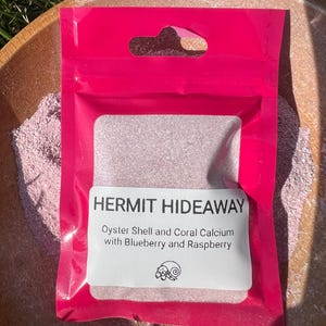May include: A bright pink, sealed plastic pouch with a hang hole, containing a white powder. The label reads "HERMIT HIDEAWAY" and lists ingredients: Oyster Shell and Coral Calcium with Blueberry and Raspberry. A small illustration of a snail is also present.