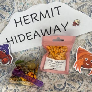 May include: A pink pouch with a resealable zipper contains a snack called "Hermit Hideaway Puff the Magic Snack" with the text "Puffed Rice, Garlic, Pepper, Peanut Oil, Honey Granules". The pouch is on a white background with the text "HERMIT HIDEAWAY" in black.
