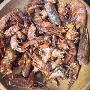 May include: A wooden bowl filled with cooked shrimp. The shrimp are a mix of orange and brown hues, with visible shells and appendages. The arrangement is dense, with many shrimp overlapping each other.