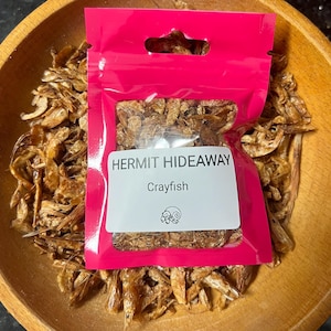May include: A pink package of dried crayfish labeled "HERMIT HIDEAWAY Crayfish" sits in a wooden bowl. The package contains small, dried, brown crayfish. The bowl is made of light brown wood.