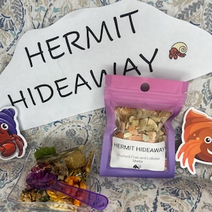 May include: A purple bag with the text "HERMIT HIDEAWAY Crushed Crab and Lobster Shells" on it. The bag contains a mixture of crushed crab and lobster shells. There are also two cartoon hermit crabs on stickers, one with a purple hat and one with a brown shell.