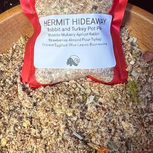May include: A red, clear plastic bag containing a mix of dried herbs and ingredients. The bag has a white label with the text "HERMIT HIDEAWAY" and ingredient information. The ingredients include rabbit, turkey, and various fruits and vegetables.
