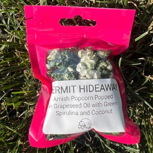 May include: A bright pink plastic bag filled with Amish popcorn. The popcorn is white and green, with the text "ERMIT HIDEAWAY" and product information on a white label. The bag is resting on green grass.