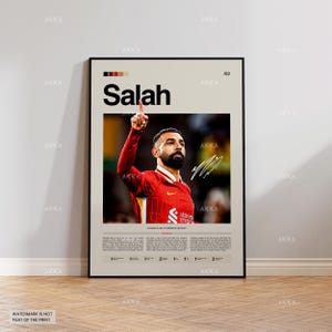 May include: A framed poster featuring the name "Salah" in large black letters. The poster includes a photograph of a soccer player in a red jersey, pointing upwards. The poster has a beige background with a black frame and a signature.