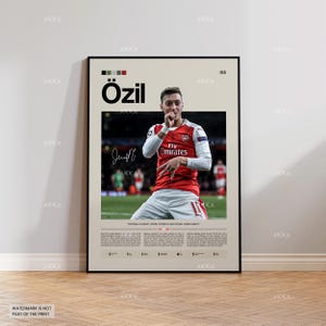 May include: A framed poster featuring a photograph of soccer player Mesut Özil in a red and white jersey. The poster includes the player's name, signature, and the text "Fly Emirates." The poster has a beige background with a black frame.