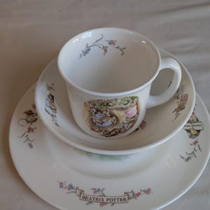 May include: A white ceramic tea set featuring a cup, saucer, and plate. The set is decorated with floral accents and a Beatrix Potter design. The cup and saucer have a small illustration of rabbits. The plate has the words "Beatrix Potter".