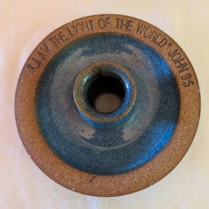 May include: A round ceramic candle holder with a textured brown outer rim and a smooth blue glazed center. The rim features the embossed text "I AM THE LIGHT OF THE WORLD" JOHN 9:5. The piece has a central circular opening for a taper candle.