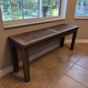 May include: A dark brown wooden bench with a slatted seat and four sturdy legs. The bench is positioned near a window, showcasing a simple, functional design. The bench measures approximately 60 inches long.