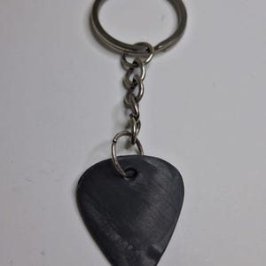 May include: A black guitar pick keychain with a silver-colored key ring and chain. The pick is teardrop-shaped and has a textured surface. The keychain is designed for guitar players.