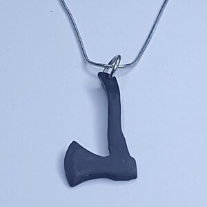 May include: A dark gray axe pendant hangs from a silver chain. The axe pendant is a solid, three-dimensional shape with a detailed blade and handle. The necklace is displayed against a light background.