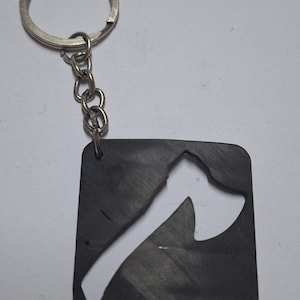 May include: A dark gray keychain featuring a square pendant with a white axe design cut out. The keychain includes a silver key ring and a short chain. The axe design is a simple, stylized illustration.