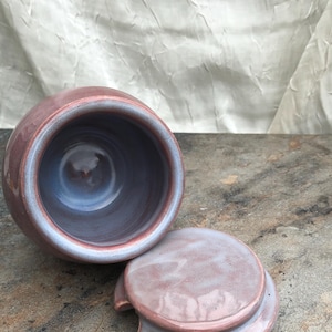 May include: A handmade ceramic jar with a lid. The jar is a dusty rose color with a blue interior and a matching lid. The lid has a small handle for easy removal. The jar is sitting on a stone surface.