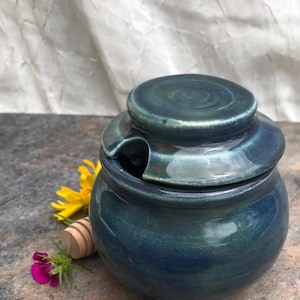 Op de afbeelding: Een handgemaakte, blauwgroene keramische honingpot met een bijpassende deksel. De pot heeft een kleine opening voor een honingdipper. Een houten honingdipper en bloemen liggen naast de pot. De pot staat op een stenen oppervlak.