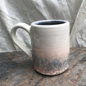 May include: A ceramic mug with a white glaze and a forest scene. The mug has a light pink and grey gradient, with a detailed forest design at the base. The handle is large and curved. The mug is suitable for hot drinks.