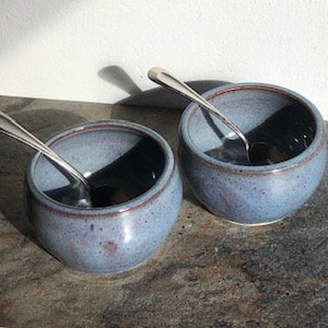 May include: Two blue ceramic bowls with a speckled glaze. The bowls are slightly rounded and have a lip around the top. Each bowl has a silver spoon inside.