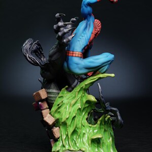 May include: A detailed figurine of Spider-Man in a dynamic pose, clad in his signature blue and red suit. He is perched atop a vibrant green, textured base, with a dark, clawed figure reaching towards him. The figurine has the Tanuki Figures logo.