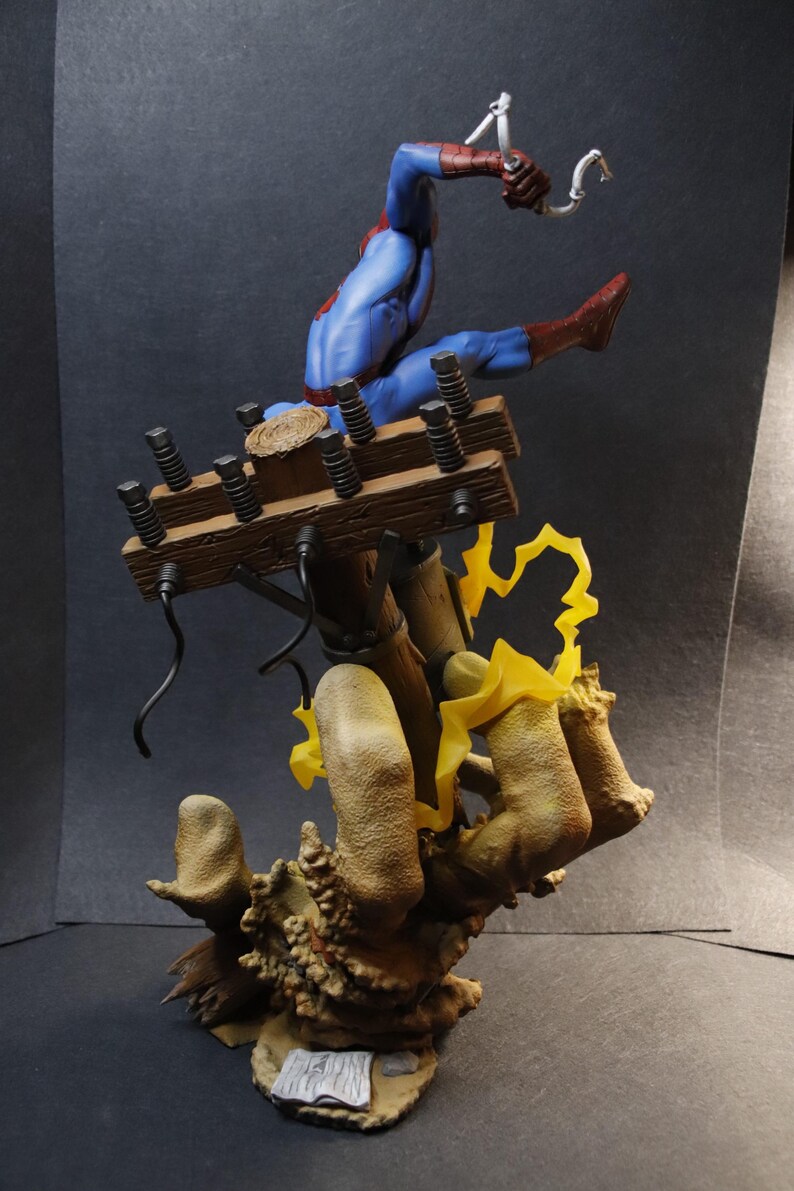May include: A detailed figurine of Spider-Man in a dynamic pose, clad in his signature blue and red suit. He is suspended mid-air, seemingly swinging from a wooden structure with electrical components. The base features a textured, organic design with yellow lightning effects.