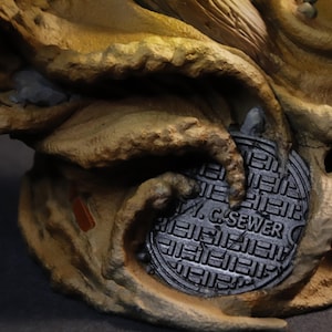 May include: A detailed sculpture featuring a textured, tan and brown organic form with a metallic gray sewer cover. The sewer cover is round and has the text "W. C. SEWER" on it. The sculpture has a dark background.