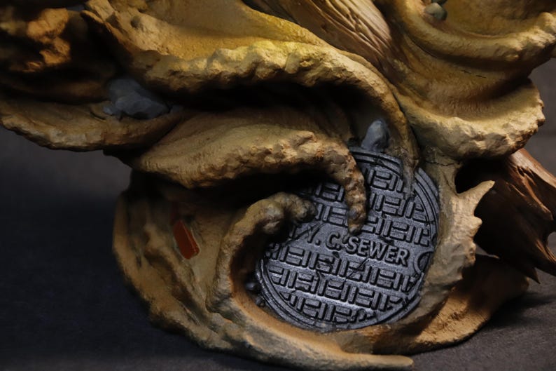 May include: A detailed sculpture featuring a textured, tan and brown organic form with a metallic gray sewer cover. The sewer cover is round and has the text "W. C. SEWER" on it. The sculpture has a dark background.