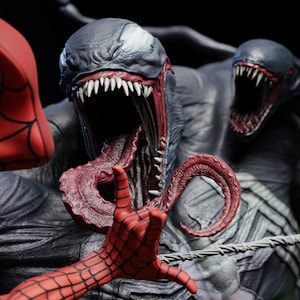May include: A detailed statue of Venom, a Marvel Comics character, with a menacing open mouth and long tongue. The statue features a red hand with a web pattern, and a second Venom head in the background. The statue is dark gray and black.