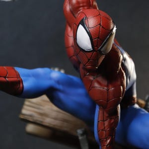 May include: A detailed figurine of Spider-Man in a dynamic pose. The figure is predominantly red and blue, with a textured pattern on the red sections. The eyes are white, and the figure appears to be mid-action, with a web strand visible.