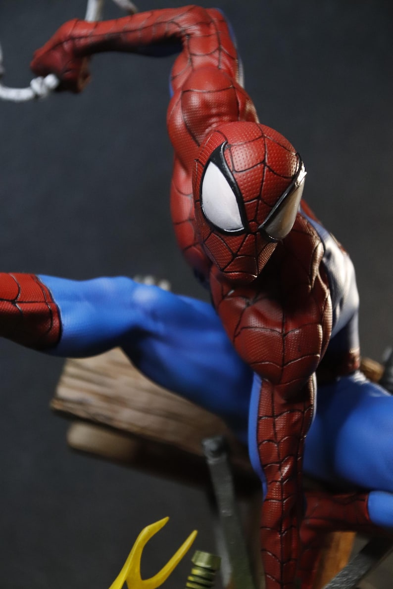 May include: A detailed figurine of Spider-Man in a dynamic pose. The figure is predominantly red and blue, with a textured pattern on the red sections. The eyes are white, and the figure appears to be mid-action, with a web strand visible.