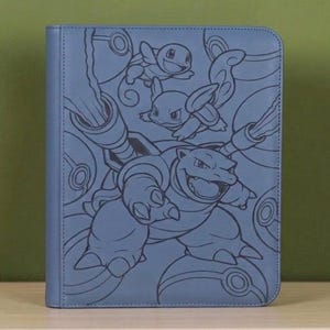 May include: A rectangular blue binder featuring a dark blue line art illustration of the Pokemon Blastoise, Wartortle, and Squirtle. The cover design includes circular patterns resembling Poke Balls integrated into the background artwork.