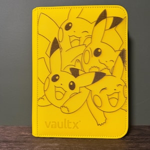 May include: Yellow card binder featuring a Pikachu design. The cover displays multiple Pikachu illustrations in a repeating pattern. The "vaultx" logo is visible at the bottom. The binder is rectangular with rounded corners.