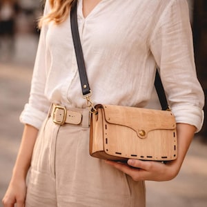 Peut inclure: Sac bandoulière en bois brun clair avec une bandoulière en cuir noir et des accessoires dorés. Le sac a une fermeture à rabat et des coutures décoratives. La personne porte une chemise blanche et un pantalon beige.