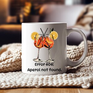 May include: White ceramic mug featuring a graphic of two Aperol Spritz cocktails with orange slices and straws. The mug has the text "Error 404: Aperol not found." printed below the graphic. The mug has a curved handle.