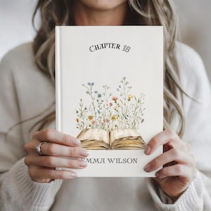 Pode incluir: Um livro de capa dura de cor creme. A capa apresenta o texto "CHAPTER 18" acima de uma ilustração de um livro aberto com flores silvestres coloridas. O nome "EMMA WILSON" é impresso na parte inferior.