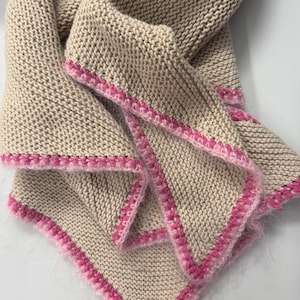Può includere: Una coperta beige lavorata a maglia con un bordo rosa. La coperta è piegata, mostrando la trama della maglia e il bordo rosa a contrasto. I bordi sono rifiniti con una delicata cucitura rosa.