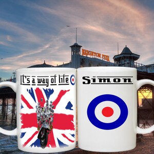 Puede incluir: Dos tazas de cerámica blanca con un diseño en cada una. La taza de la izquierda tiene una bandera de la Union Jack con una scooter y el texto "It's a way of life" y un blanco, rojo y azul objetivo. La taza de la derecha tiene el nombre "Simon" y un blanco, rojo y azul objetivo.