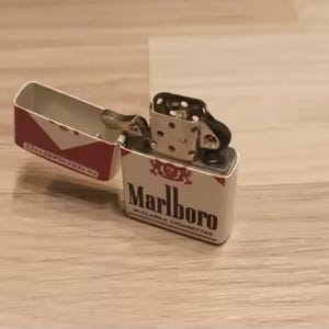 Puede incluir: Un encendedor de metal con tapa abatible en acabado blanco y rojo que presenta el logotipo y el texto de Marlboro. El encendedor est&aacute; abierto, revelando la chimenea interna plateada y el mecanismo de la rueda. El exterior muestra la marca y el texto 20 Class A Cigarettes.